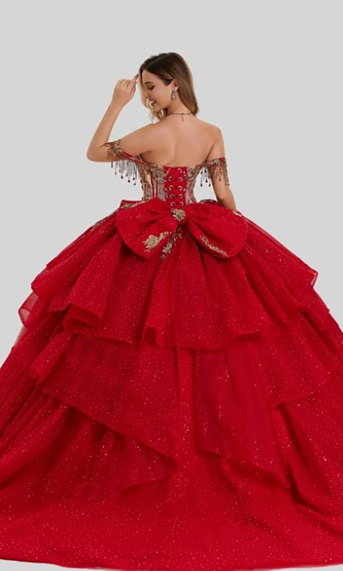 A woman stands against a plain gray background, shown from the back, wearing the Quinceanera Dress 26417 by Stella Couture—an off-shoulder, layered red ball gown adorned with a large bow and gold detailing, featuring a gold-embellished corset bodice.
